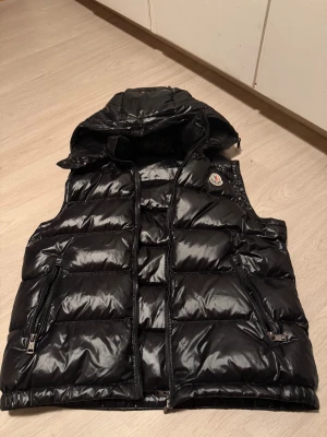 Svart dunväst från Moncler med avtagbar huva - Äkta moncler väst glansig med avtagbar luva använd fåtal gånger köptes av en polare till mig som köpte den ny för 12000kr på Careofcarl. Kvitto finns inte tyvär. Storlek 2 