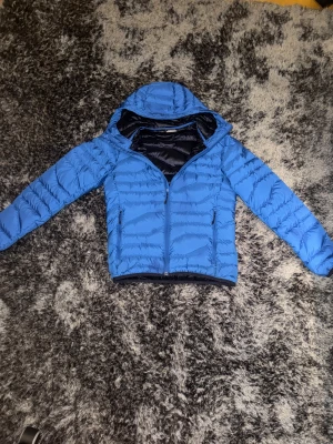 Blå dunjacka från J.Lindeberg - Snygg blå dunjacka från J.Lindeberg med huva och dragkedja framtill. Jackan har quiltad design, två sidofickor med dragkedja och logga på bröstet. Perfekt för kalla dagar och har en modern, sportig look. Ny pris 2300kr
