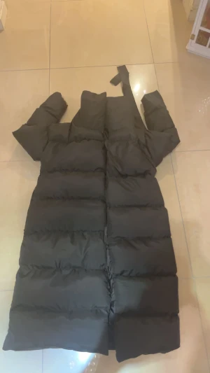 Svart lång pufferjacka med huva - Säljer en svart lång pufferjacka med huva och breda, quiltade sektioner. Jackan har en rak och oversized passform, perfekt för kalla dagar. Den är utrustad med dragkedja framtill och har långa ärmar. Enkel och stilren design som håller dig varm hela vintern.