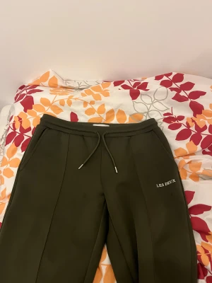 Mörkgröna mjukisbyxor Les Deux - Snygga mörkgröna mjukisbyxor från Les Deux med stilren design och broderad logga på benet. Byxorna har snörning i midjan och raka ben med markerade sömmar framtill. Perfekta för chill eller streetwear. Materialet är mjukt och skönt.