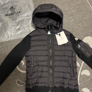 Svart Moncler pufferjacka med huva - Snygg svart pufferjacka från Moncler med stickade ärmar och quiltad kropp. Jackan har huva, dragkedja framtill och Moncler-logga på ärmen. Fickor med tryckknappar och ribbad mudd vid midja och ärmslut. Perfekt för dig som vill ha en cool och exklusiv look.