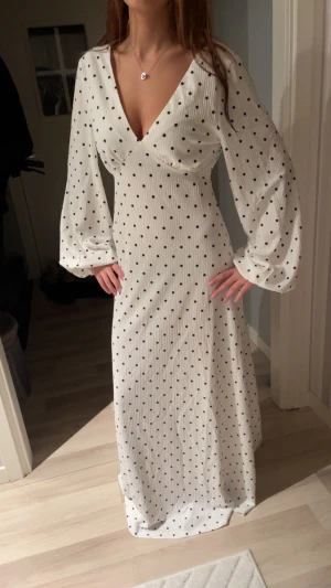 Vit maxiklänning med svarta prickar NA-KD - Superfin vit maxiklänning från NA-KD med svarta prickar, ballongärmar och djup v-ringning. Klänningen har öppen rygg med knytband i nacken och ett flowy fall. Perfekt för dig som vill sticka ut med en elegant och trendig look.