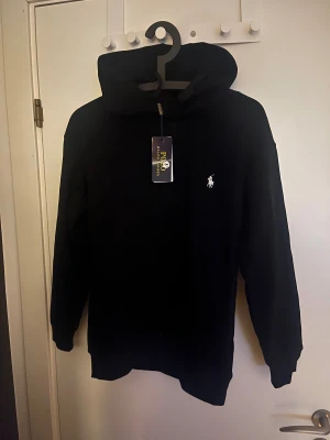 Svart hoodie från Polo Ralph Lauren - Svart hoodie från Polo Ralph Lauren med klassisk vit logobrodyr på bröstet. Tröjan har huva, långa ärmar och ribbade muddar. Perfekt för dig som gillar stilrena och tidlösa plagg med premiumkänsla.