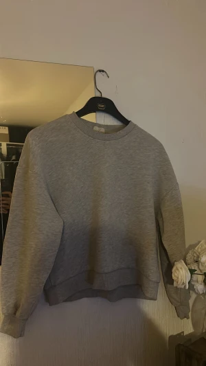 Grå sweatshirt med rund hals - En enkel och stilren grå sweatshirt med rund hals och ribbade muddar vid ärmslut och nederkant. Tröjan har en loose passform och är långärmad, perfekt för chill dagar. Materialet känns mjukt och bekvämt, troligen bomull eller polyester.