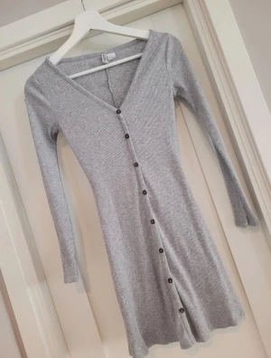 Grå ribbad klänning från Divided XS - Superfin grå ribbad klänning från Divided i storlek XS. Klänningen har v-ringning, långa ärmar och knappar hela vägen framtill. Den är figurnära och kort i modellen, perfekt för en stilren och enkel look.