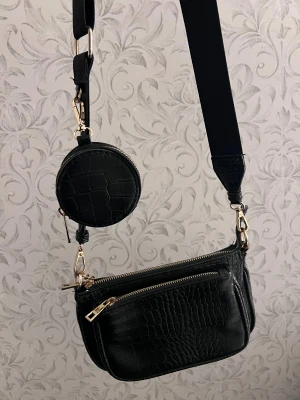 Svart axelväska med croco-mönster - Snygg svart axelväska med croco-mönster och guldfärgade detaljer. Väskan har två fack med dragkedja och en rund liten coin pouch som hänger på den breda axelremmen. Hör av dig för frågor🩷