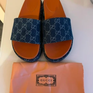 Gucci sandaler i blå denim - Snygga sandaler från Gucci med bred rem i blå denim och klassiskt GG-mönster. Sulan är brun och har Guccis logga undertill. Perfekt för sommaren och riktigt trendiga med öppen tå och platt sula. Kommer med original dustbag.
