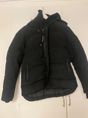 Svart pufferjacka från Canada Goose - Svart pufferjacka från Canada Goose med huva och diskret logopatch på ärmen. Jackan har dragkedja och tryckknappar framtill, två sidofickor med dragkedja och quiltad design. Perfekt för kalla dagar och har en clean, modern look. Använd endast en vinter så riktigt bra sick. Kommer med sin påse. Kan gå ner i pris!