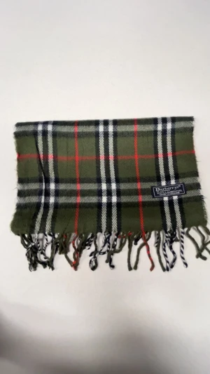 Rutig grön halsduk från Burberrys - Snygg rutig halsduk från Burberrys i mörkgrön ton med inslag av vitt, svart och rött. Gjord i 100% lammull och har klassiska fransar i ändarna. Perfekt accessoar för att lyfta din outfit med lite brittisk vibe.