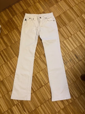 Rock & Republic vita bootcut jeans - Snygga vita jeans från Rock & Republic Victoria Beckham-kollektionen med bootcut passform. Jeansen har glittriga kronbroderier på bakfickorna och klassisk femficksdesign. Tillverkade i 99% bomull och 1% elastan för skön stretch.