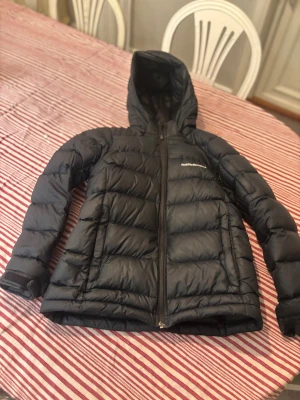 Svart Peak Performance dunjacka 160 - Svart dunjacka från Peak Performance med huva och dragkedja framtill. Jackan har quiltad design och logga på bröstet. Materialet är syntet och dun, perfekt för kalla dagar. Snygg och sportig look med långärmade ärmar och justerbara muddar. Påse ingår😃Priset går självklart att diskutera. Passar på både tjejer och killar.