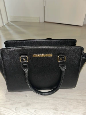 Svart handväska från Michael Kors - Stilren svart handväska från Michael Kors med guldfärgade detaljer och logga framtill. Väskan har både handtag och axelrem, samt ett rymligt fack med dragkedja. Tillverkad i strukturerat skinn med klassisk, fyrkantig form. Perfekt för dig som gillar snygga och praktiska accessoarer.