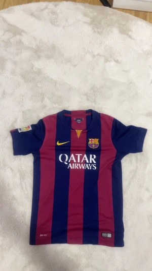 FC Barcelona matchtröja Nike 2014 (äkta)  - Säljer en äkta FC Barcelona matchtröja från säsongen 2014 med Qatar Airways-tryck, broderat klubbmärke och LFP-patch på ärmen. Tröjan är kortärmad, har breda blå och vinröda ränder och är tillverkad i Dri-FIT-material för bästa komfort.