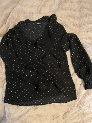 Svart prickig blus från Gina Tricot - Säljer en svart blus från Gina Tricot i storlek 34 med vita prickar och volangdetaljer längs halsringningen och ärmarna. Blusen har långärmade ärmar med knappar vid ärmslut och en omlott-look framtill. Perfekt för dig som gillar trendiga mönster och snygga detaljer.