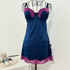 Marinblå kort negligé med spets - Snygg marinblå negligé med lila spetsdetaljer vid byst och nederkant. Klänningen har axelband, öppen rygg och en liten svart rosett med pärla vid slitsen. Storlek: 75C, passar byst 85-90cm, underbyst 73-75cm.