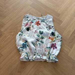 Blommig linnetopp från Stradivarius - Säljer en söt linnetopp från Stradivarius i vit med färgglatt blommigt mönster. Toppen är ärmlös och har volangkant nedtill samt en liten knapp i nacken. Perfekt för sommarens alla häng och picknickar!