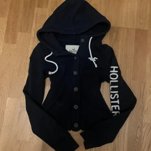 VintageMörkblå hoodie från Hollister  - Snygg mörkblå hoodie från Hollister med vit broderad logga på bröstet och stort Hollister-tryck längs ena ärmen. Tröjan har knappar hela vägen och långa ärmar🔵| skriv vid funderingar priset kan självklart diskuteras!!