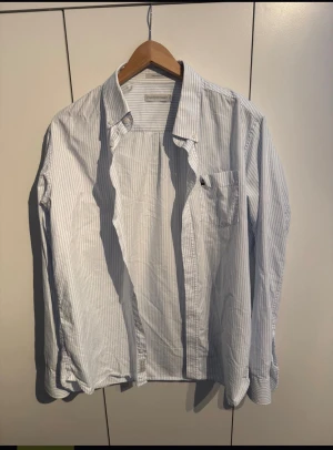 Oxford skjorta Peak Performance - Snygg skjorta från Peak Performance i vitt och ljusblått med tunna vertikala ränder. Klassisk krage, bröstficka och knappar framtill. Långärmad och tillverkad i mjuk bomull, perfekt för en clean och fräsch look.