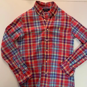 Rutig röd skjorta från Polo Ralph Lauren - Snygg rutig skjorta från Polo Ralph Lauren i slim fit. Skjortan har en klassisk krage, långa ärmar och knappar framtill. Färgerna är röd, blå, gul och vit med broderad logga på bröstet. Perfekt för dig som gillar färgstarka och stilrena plagg.