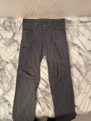 Grå cargopants från Haglöfs, M - Säljer ett par grå cargopants från Haglöfs i storlek M. Byxorna har flera praktiska fickor, bland annat med dragkedja på benet, och är tillverkade i slitstarkt syntetmaterial. Perfekta för dig som gillar outdoor och funktionella kläder.