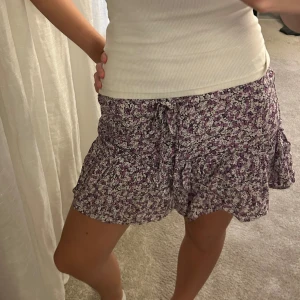 Lila blommig kortkjol med volang - Supersöt kortkjol med lila och vita blommor, volangkant och knytband i midjan. Kjolen har en omlott-design som ger en skön och luftig känsla. Perfekt för dig som gillar romantiska och lekfulla detaljer i din stil.