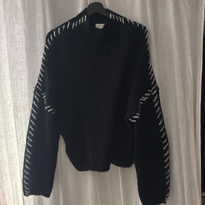 Svart stickad tröja med snörning från Vila - Cool svart stickad tröja från Vila i storlek L. Tröjan har grov snörning i vitt längs axlar och ärmar som ger en unik look. Modellen är loose fit med långärmade ärmar och hög hals. Perfekt för dig som gillar statement-plagg och vill sticka ut.