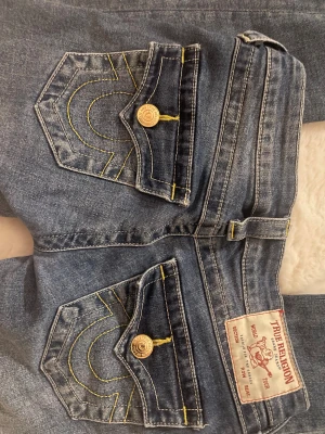 True Religion Joey Low Rise Flare Jeans - Snygga blå jeans från True Religion, modell Joey Low Rise Flare. Byxorna har låg midja, utsvängda ben och klassiska fickor med gul kontrastsöm och dekorativa knappar. Tillverkade i jeansmaterial med coola detaljer och ikonisk logga bak. Passar 152 har sytt upp dem