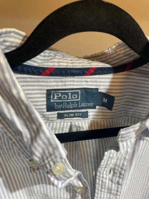 Randig skjorta Polo Ralph Lauren M - Snygg slim fit skjorta från Polo Ralph Lauren i vitt och blått med tunna ränder. Klassisk krage, knappar framtill och broderad logga på bröstet. Långärmad och tillverkad i mjuk bomull. Perfekt för dig som gillar stilrena och tidlösa plagg.