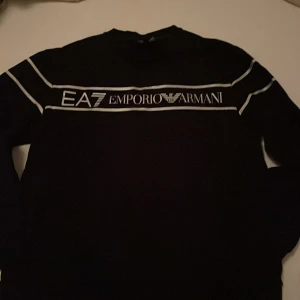Svart EA7 Emporio Armani sweatshirt - Svart långärmad sweatshirt från EA7 Emporio Armani med vit logga och vita linjer över bröstet och axlarna. Tröjan har en klassisk rund hals och är tillverkad i mjuk bomull för skön komfort. Perfekt för dig som gillar stilrena och sportiga märkesplagg.