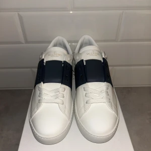 Valentino Garavani Open sneakers vit/blå - Snygga Valentino Garavani Open sneakers i vitt skinn med bred marinblå rem över mitten. Klassisk rund tå, vita snören och coola nitar runt hälen. Sulan är vit med rutmönster undertill. Perfekta för dig som gillar stilrena och exklusiva sneakers.