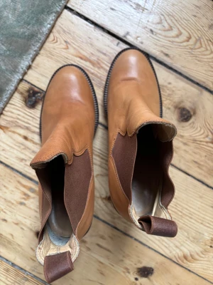 Acne Studios bruna chelsea boots - Snygga bruna chelsea boots från Acne Studios i äkta skinn. Boots med rund tå, bred klack och elastiska paneler i mörkbrunt på sidorna. Tillverkade i Italien, klassisk siluett och robust träklack som ger extra höjd. Perfekt för dig som gillar stilrena och tidlösa skor.
