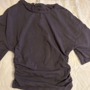 Mörkgrå croppad topp med snörning - Säljer en mörkgrå croppad topp med korta ärmar och snygg snörning på ena sidan. Toppen har en avslappnad passform och är tillverkad i mjuk bomull. Perfekt för dig som gillar enkla men coola plagg med en twist.