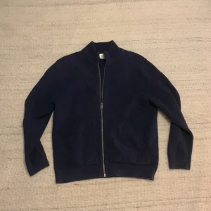 Mörkblå stickad kofta med dragkedja - Säljer en mörkblå stickad kofta med hel dragkedja framtill och två praktiska fickor. Koftan har en enkel, stilren design med ribbad struktur och långärmade ärmar. Perfekt för att hålla värmen under kyliga dagar. Lite mindre än xs.😄