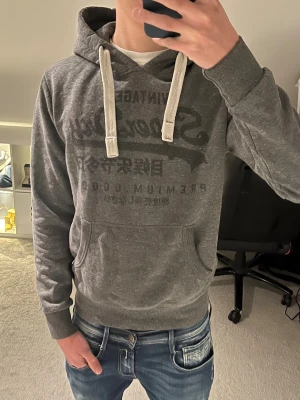 Grå hoodie från Superdry  - Riktigt fet mörkgrå hoodie från Superdry med stort vintage-tryck på bröstet och vita snören i huvan. Riktigt bra passform, i storlek M men sitter som S/M.