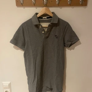 Grå piké från Abercrombie & Fitch - Snygg grå pikétröja från Abercrombie & Fitch med klassisk krage och korta ärmar. Tröjan har knappar vid halsen och en diskret broderad logga på bröstet. Perfekt för en chill och stilren look.