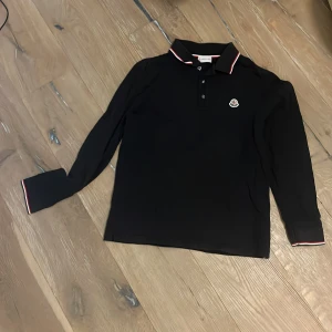 Blå långärmad pikétröja Moncler - Blå långärmad pikétröja från Moncler med klassisk krage och knäppning framtill. Tröjan har diskret Moncler-logga på bröstet och snygga rödvita detaljer på krage och ärmslut. Perfekt för dig som gillar stilrena och sportiga plagg.