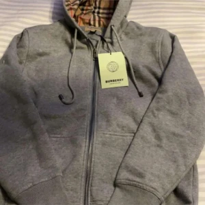 Grå hoodiejacka från Burberry - Grå hoodiejacka från Burberry med klassiskt rutigt foder i beige, svart och rött. Jackan har dragkedja framtill, snörning i huvan och ribbade muddar. Perfekt för dig som vill ha en stilren men ändå lyxig look. Snabb affär billigare pris