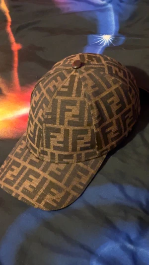 Brun fendi keps - Säljer en snygg keps från Fendi med det klassiska FF-logomönstret i brunt och svart. Kepsen har justerbar rem baktill i mörkbrunt läder och Fendi-logga på insidan. Perfekt accessoar för att lyfta din streetwear-look. I perfekt skick och priset kan absolut diskuteras vid snabb affär.