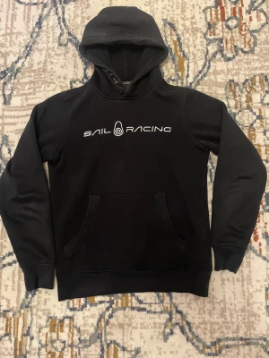 Svart hoodie från Sail Racing XS - Svart hoodie från Sail Racing i storlek XS med vit logotyp på bröstet. Tröjan har huva, magficka och långa ärmar. Jag har haft den ett tag men den funkar att använda, den har lite små tecken på användning men inget som syns jättemycket.