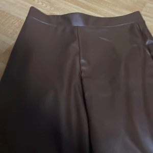 Bruna vida byxor i skinnimitation - Snygga bruna byxor i skinnimitation med vida ben och hög midja. Byxorna har en slät finish och är perfekta för dig som vill sticka ut med en trendig look. Passar till många olika outfits och ger en cool vibe. Storlek 146