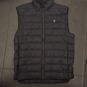 Blå dunväst med ståkrage - Mörkblå dunväst med horisontella quiltade sömmar och ståkrage. Västen har en dragkedja framtill och en liten guldfärgad logga på bröstet. Perfekt för lager-på-lager och enkel att matcha med olika outfits. Nypris 2300.