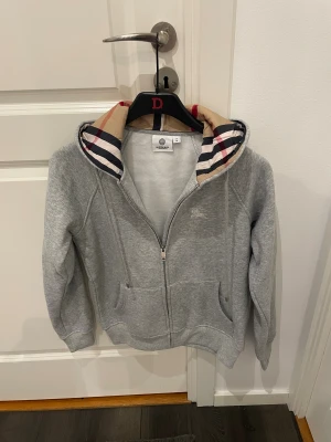 Grå Burberry hoodie med dragkedja - Snygg grå hoodie från Burberry med klassiskt rutigt mönster i huvan. Tröjan har dragkedja framtill, två fickor och långa ärmar. Materialet är mjuk bomull och polyester, perfekt för chill dagar. Ikonisk Burberry-logga broderad på bröstet. Passar storlek S kanske Xs