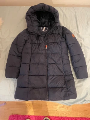 Mörkblå  lång pufferjacka Save The Duck - Säljer en mörkblå, lång pufferjacka från Save The Duck med huva och snygg quiltad design. Jackan har dragkedja framtill och ett orange logomärke på ärmen. Perfekt för kalla dagar och har en clean, modern look.