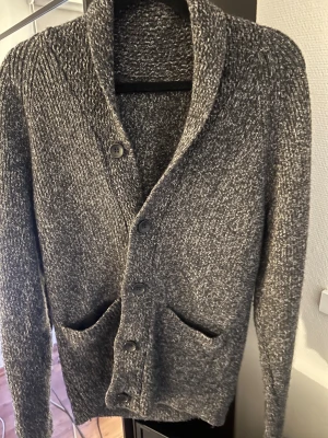 Grå stickad kofta med knappar - Modellen är 184 cm lång och väger 72 kg‼️Mysig gråmelerad stickad kofta med grov struktur och fyra svarta knappar framtill. Koftan har två praktiska fickor och en bred krage som ger en chill och avslappnad vibe. Perfekt att slänga på över en t-shirt när det är kyligt ute.