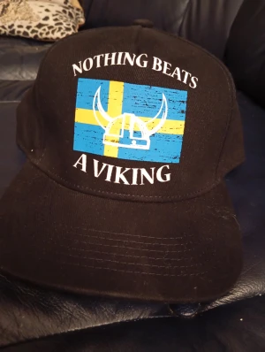 Svart keps med svensk viking-tryck - Cool svart keps med tryck av svensk flagga och vikingahjälm på framsidan. Texten 'Nothing beats a Viking' ger den extra attityd. Perfekt för dig som vill visa din svenska stolthet och vikinga-vibe. Justerbar baktill för skön passform.