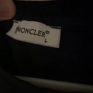 Svart Moncler t-shirt med logga - Svart t-shirt från Moncler i storlek L med klassisk rund hals och korta ärmar. På bröstet sitter Monclers ikoniska logga i vitt och rött. T-shirten är gjord i mjuk bomull och har en clean, stilren look som funkar till allt. Den passar bättre Som storlek M