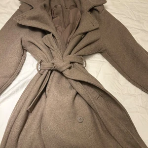 Beige omlott kappa med bälte - Snygg beige kappa med omlott-design och bälte i midjan. Kappan har stora slag, knappar framtill och långa ärmar med dekorativ knappdetalj vid mudden. Materialet är mjukt och känns lyxigt, perfekt för dig som vill ha en clean och stilren look.