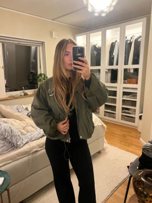 Olivgrön bomberjacka - Säljer en olivgrön bomberjacka med oversized passform och coola detaljer som snörning i nederkant, stora fickor med knappar och dragkedja framtill. Jackan har lång ärm och en snygg krage med metallspänne. 