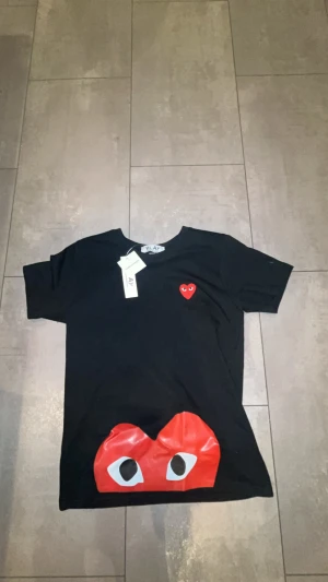 Svart Comme des Garçons Play t-shirt - Svart t-shirt från Comme des Garçons Play med ikoniskt rött hjärta med ögon broderat på bröstet och stort tryck med röda ögon längst ner. Klassisk rund hals och korta ärmar. Skön bomullskvalitet och snygg passform.
