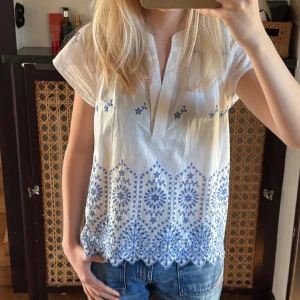 Vit blus med blå broderi Holly & White - Supersöt vit blus från Holly & White med blått broderat mönster och v-ringad hals. Blusen har korta ärmar och ett detaljerat broderi längs nederkanten och upptill. Perfekt till jeans eller kjol för en fräsch look i sommar. Lapp kvar!